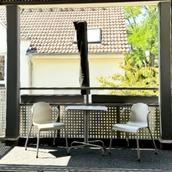 Überdachter Balkon mit kleinem Bistrotisch und zwei weißen Stühlen, geschlossenem Sonnenschirm sowie Blick auf ein Nachbarhaus mit Ziegeldach und Baum