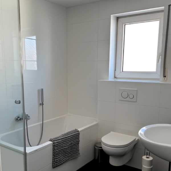Modernes, helles Badezimmer mit weißen Fliesen, Badewanne mit Glasabtrennung und Handbrause, wandhängendem WC, Waschbecken und Fenster mit Tageslicht